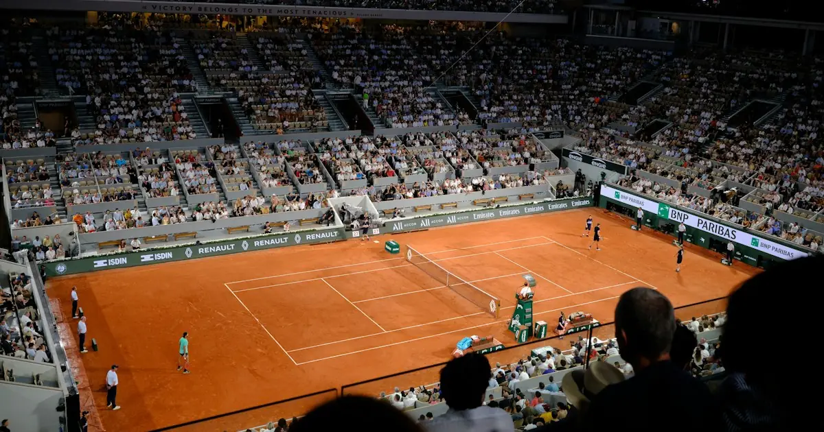 A floodlit clay tennis court in a full stadium at night